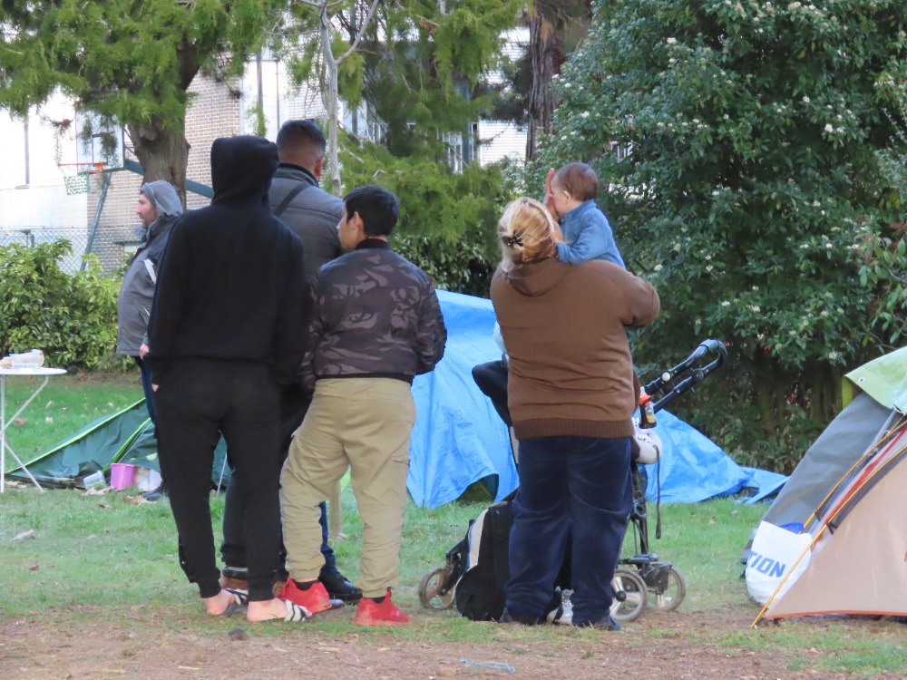 Un groupe de personne, adultes comme enfants, debout devant des tentes, plantées dans un parc.